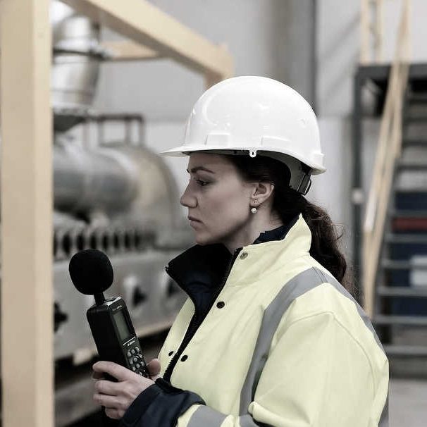 A surveyor in a hard hat holding a sound level meter in an industrial plant